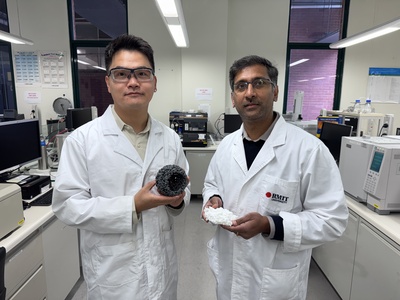 Dr Derek Hao and Associate Professor Ravichandar Babarao stand in an RMIT laboratory surrounded by scientific instruments. Dr Hao holds a grey, porous spherical model of the low-cost material developed in the study, while Associate Professor Babarao holds a white sample of the precursor material.